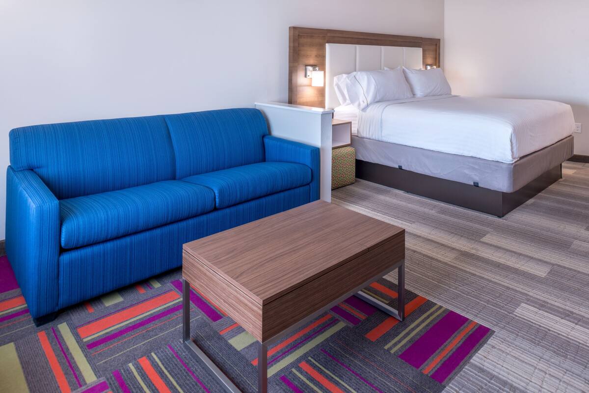 A hotel room with a blue sofa, a wooden coffee table, a bed with white bedding, and a colorful striped carpet.