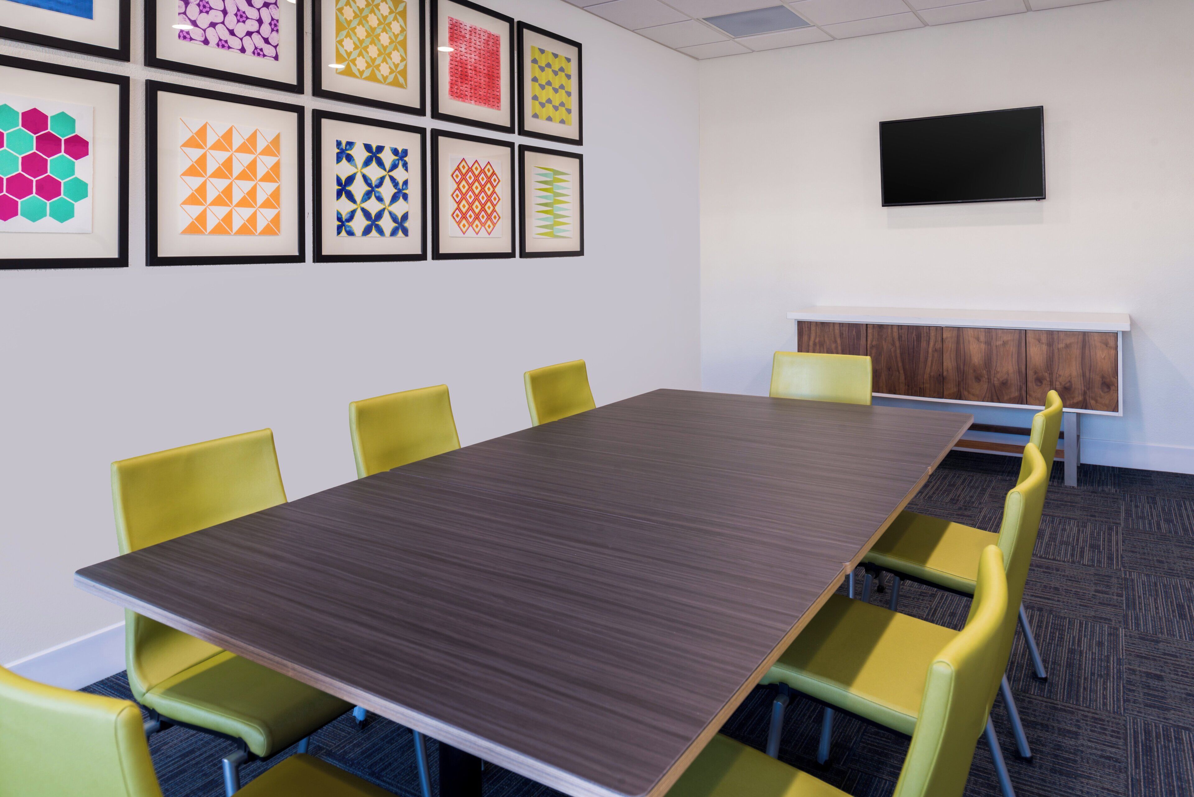 A conference room with a long table and yellow chairs, a flat-screen TV on the wall, and framed geometric patterns on the wall.