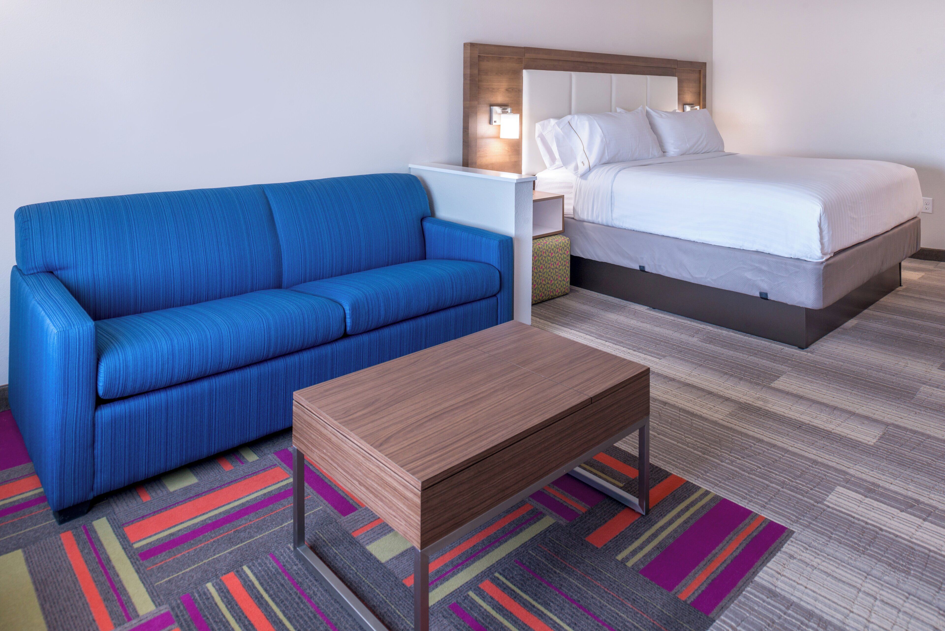 A hotel room with a blue sofa, a wooden side table, a bed with white bedding, and a colorful striped carpet.