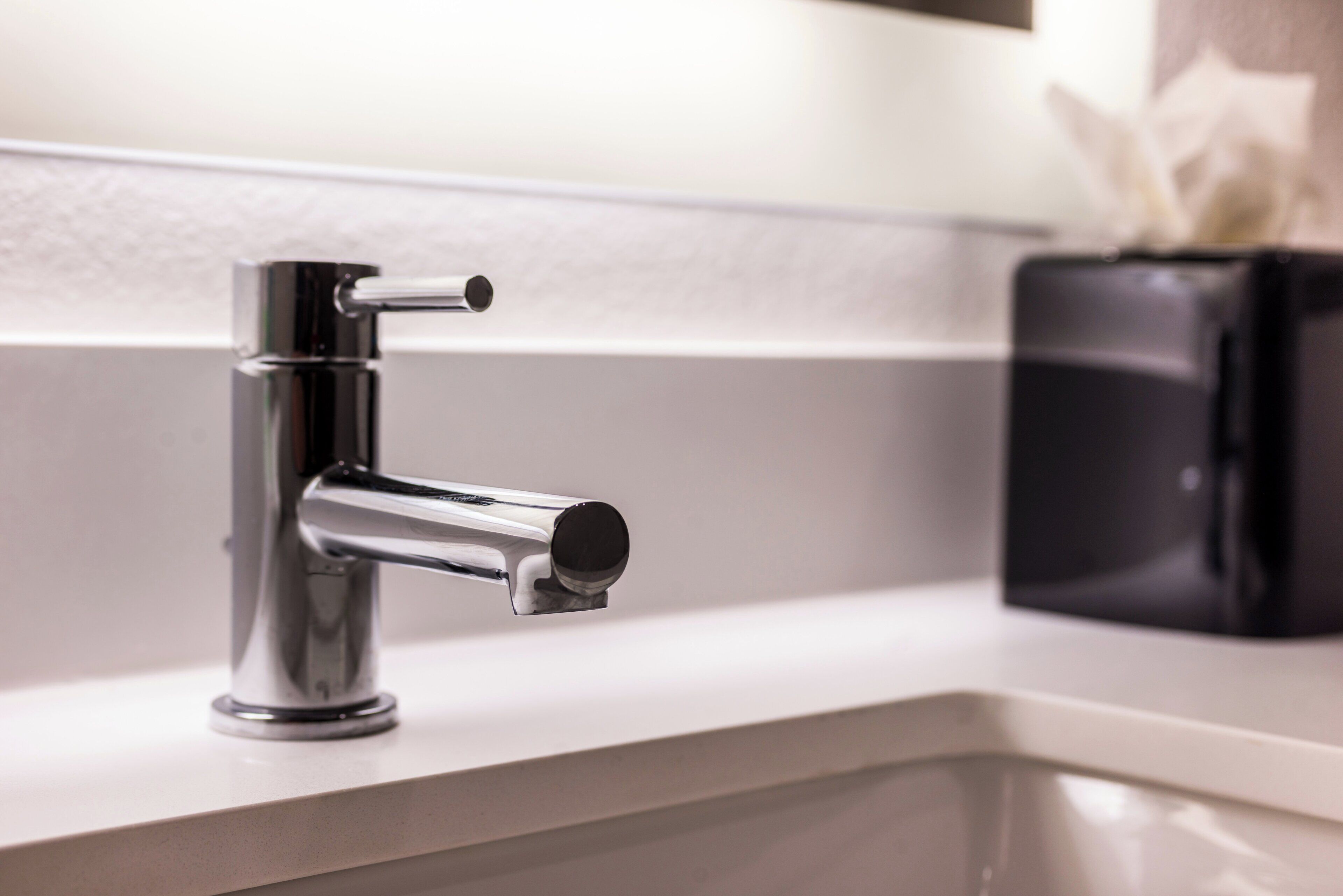 A modern bathroom with a chrome faucet, a white countertop, and a black soap dispenser.