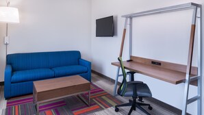 A room with a blue sofa, a wooden desk, a black office chair, a colorful striped rug, and a mounted TV on the wall. - Holiday Inn Express & Suites Fort Worth West by IHG (Fort Worth)
