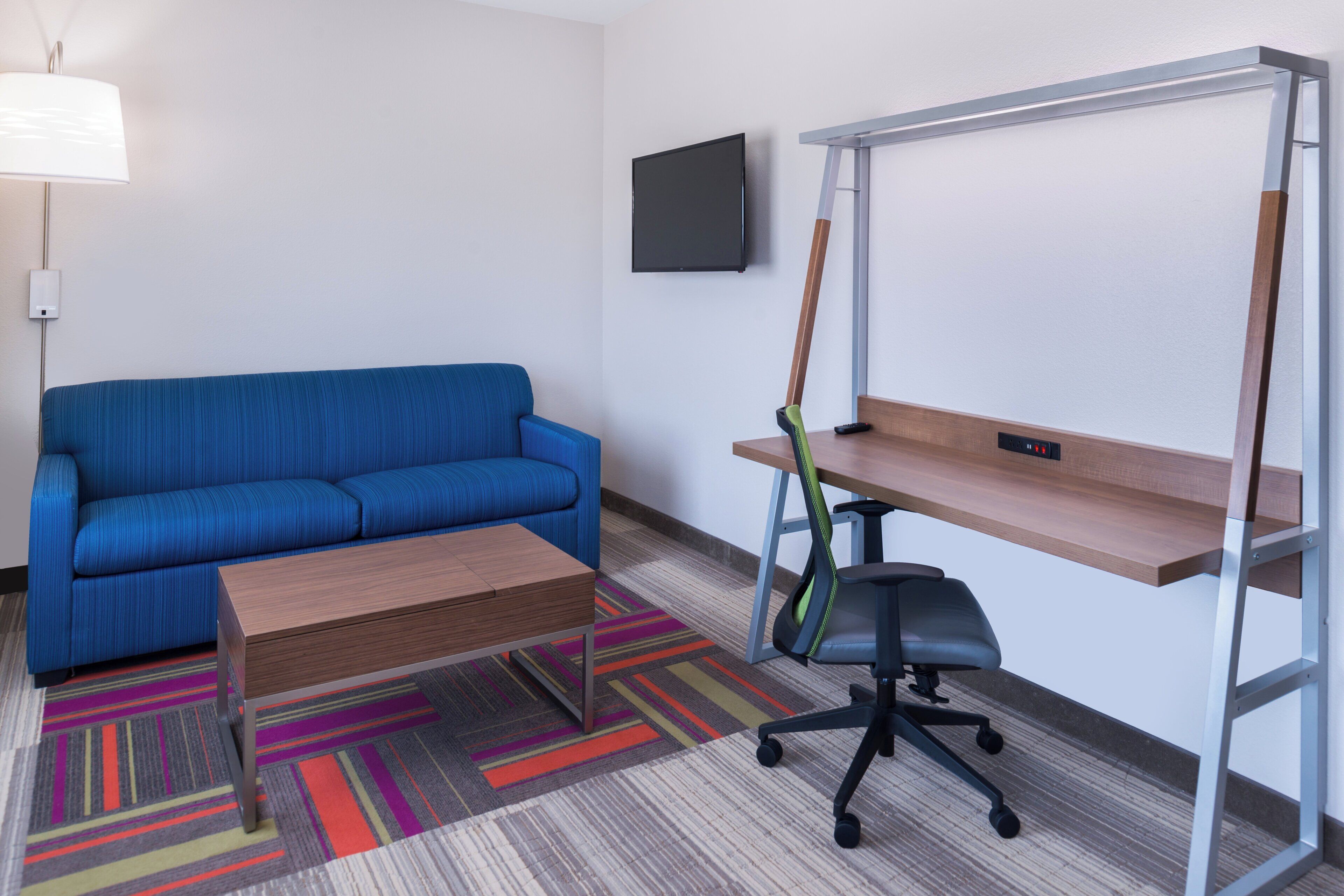 A hotel room with a blue sofa, a wooden coffee table, a desk with a chair, a TV mounted on the wall, and a colorful carpet.