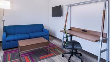A hotel room with a blue sofa, a wooden coffee table, a desk with a chair, a TV mounted on the wall, and a colorful carpet.