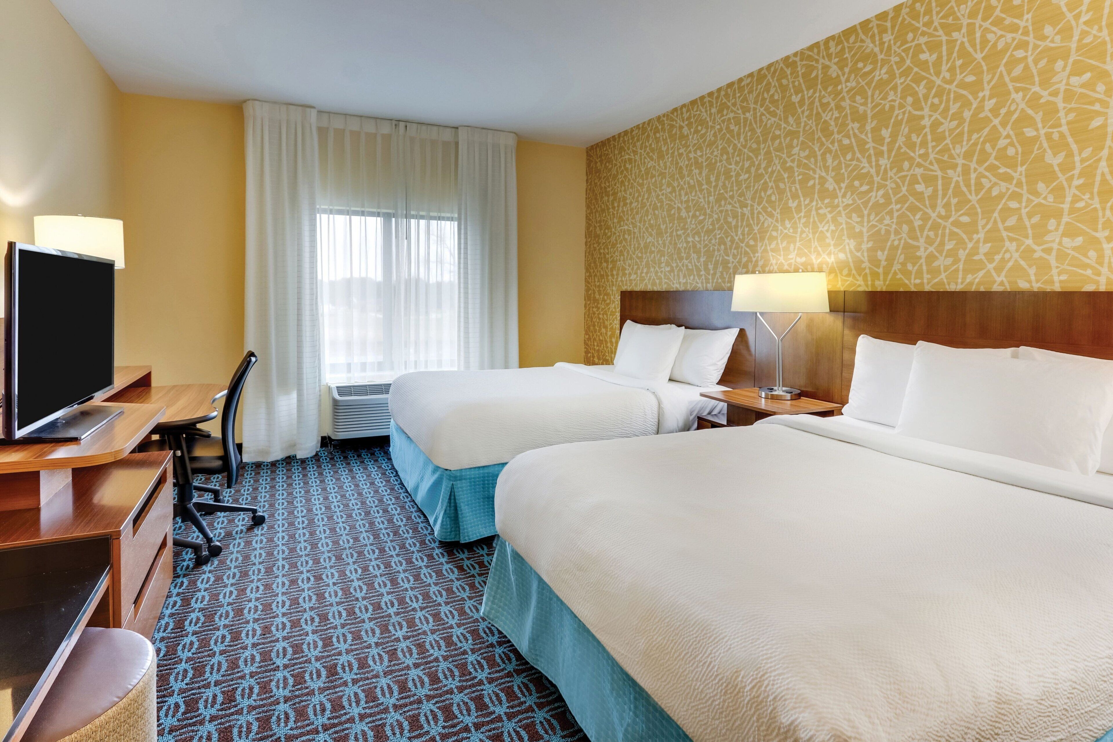 A hotel room with two beds, a desk with a television, a chair, and a patterned carpet.