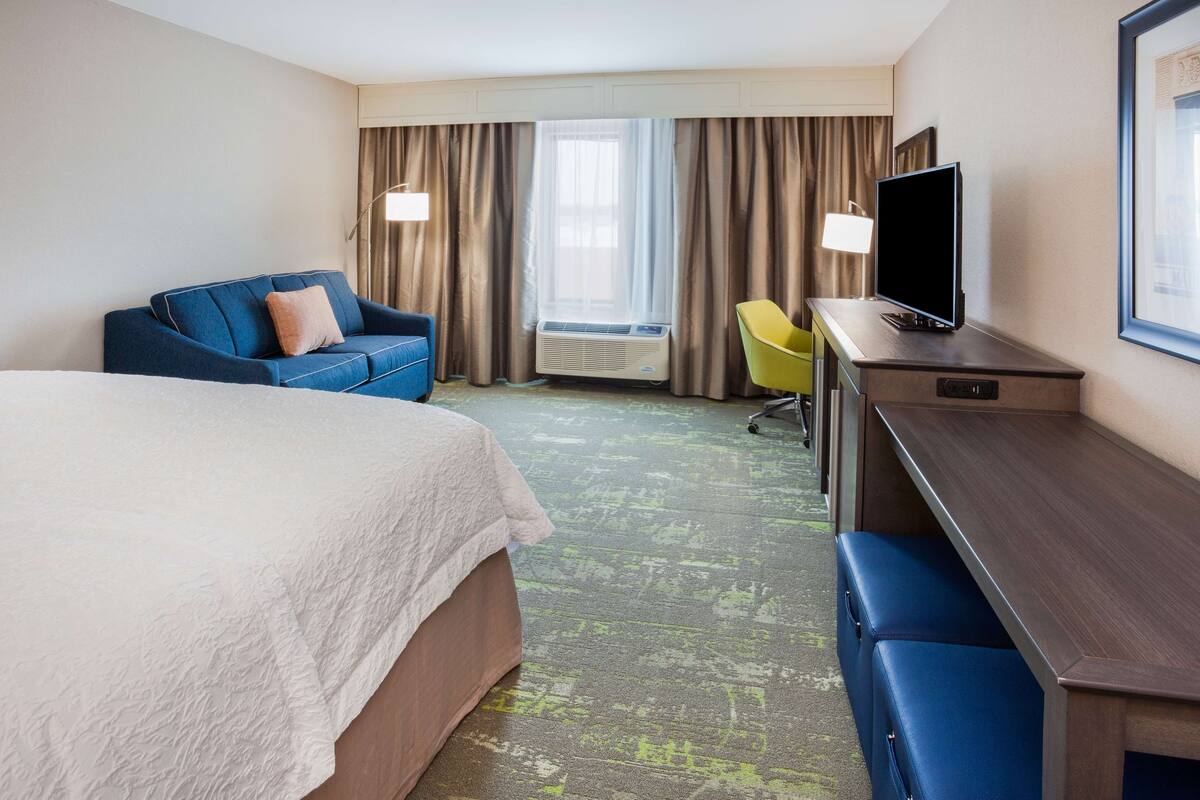 A hotel room with a bed, a blue sofa, a desk with a TV, and a window with curtains.