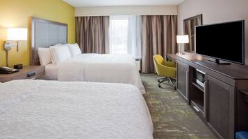 A hotel room with two beds, a desk with a flat-screen TV, a chair, and a nightstand with a lamp and phone.