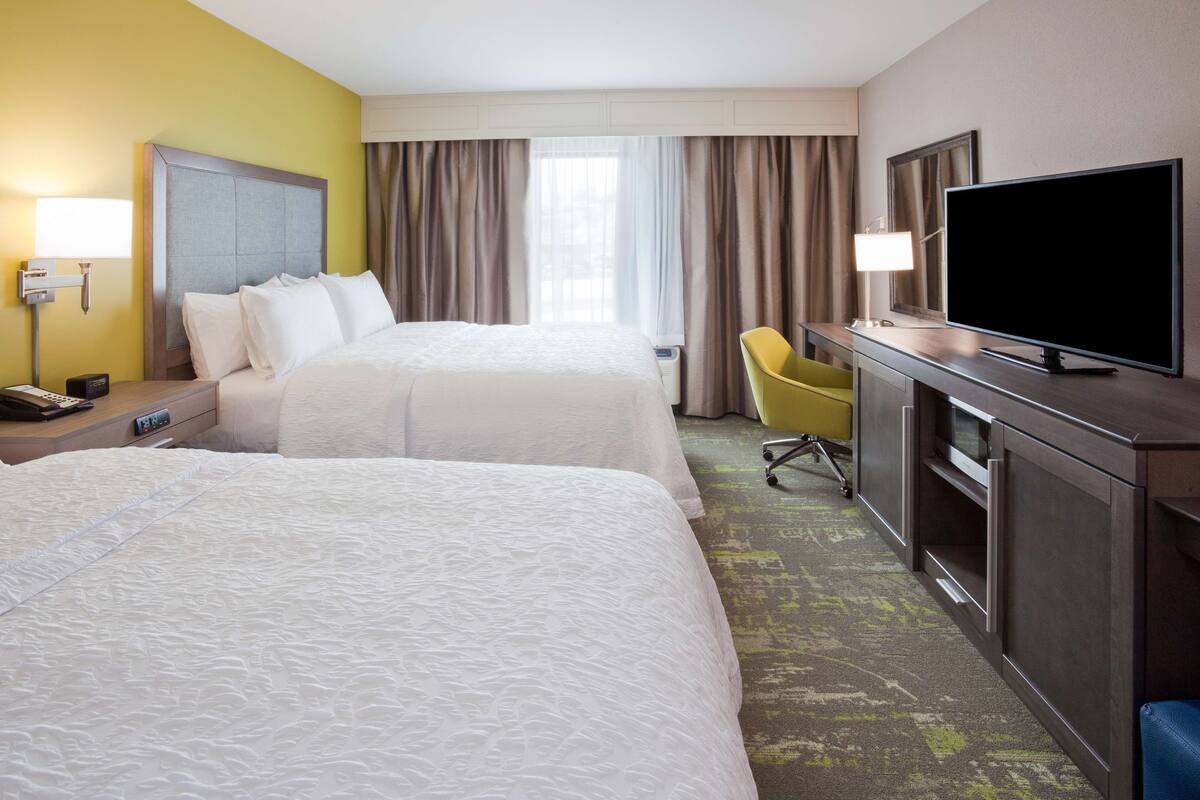 A hotel room with two beds, a desk with a flat-screen TV, a chair, and a nightstand with a lamp and phone.