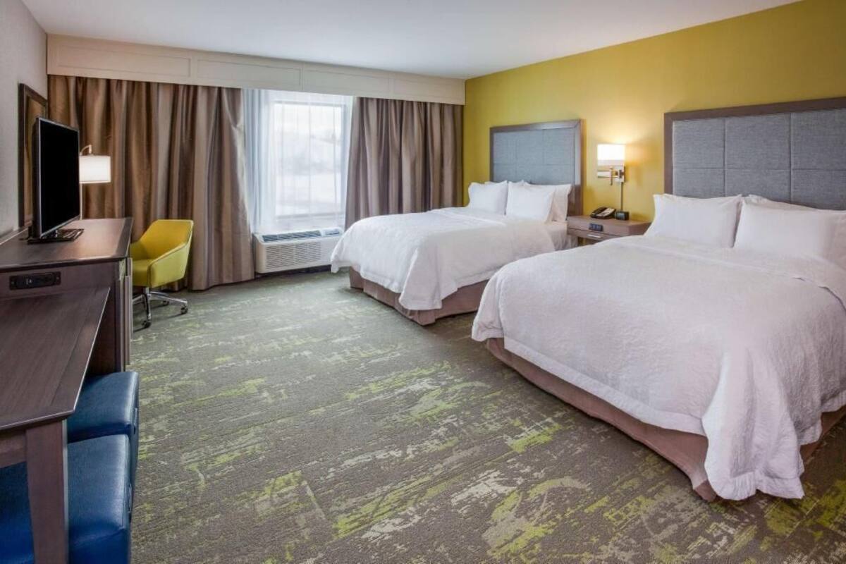 A hotel room with two beds, a desk with a chair, a TV, and a window with curtains.