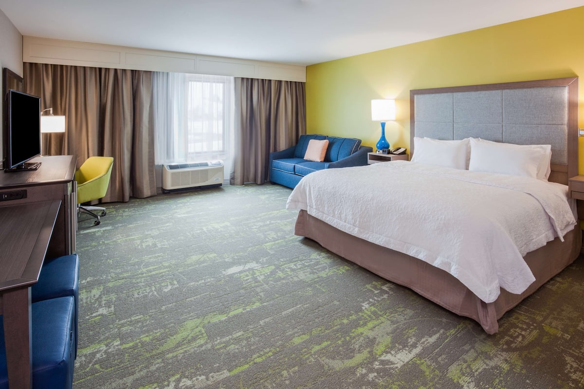 A hotel room with a large bed, a desk with a computer, a chair, a sofa, and a TV.