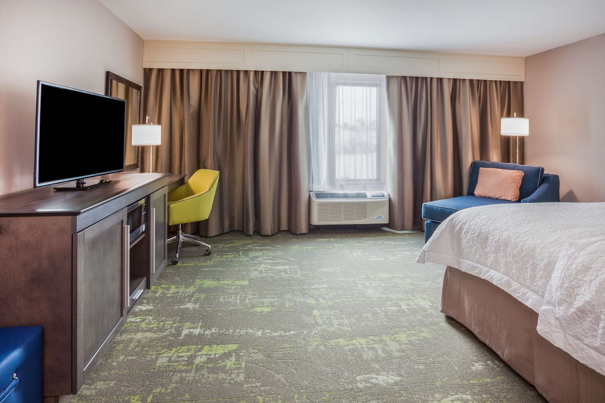 A hotel room with a bed, a desk with a TV, a chair, and a window with curtains.