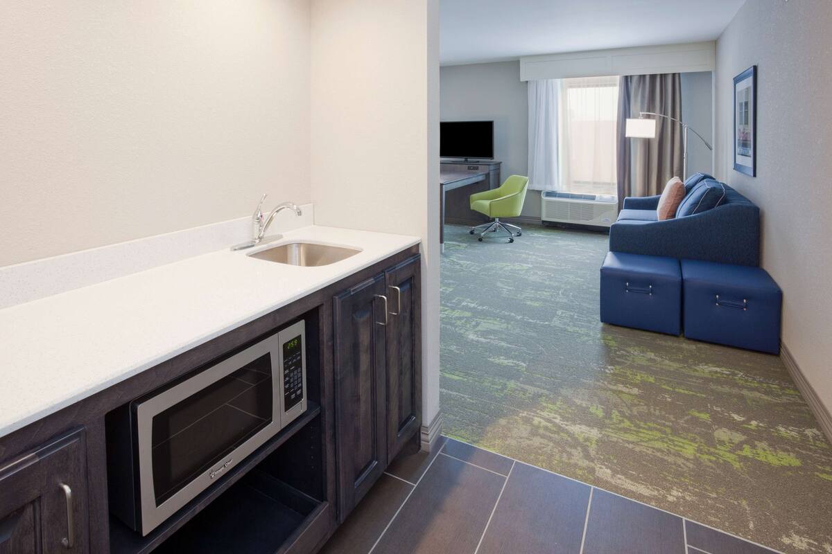 A modern hotel room with a kitchenette, a sofa, and a large window.
