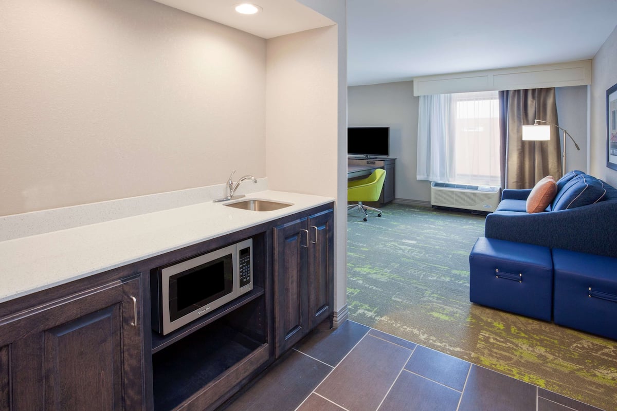 A modern kitchen with a built-in microwave, a sink, and dark wooden cabinets. There is a blue sofa with cushions in the living area, a flat-screen TV, and a window with curtains.