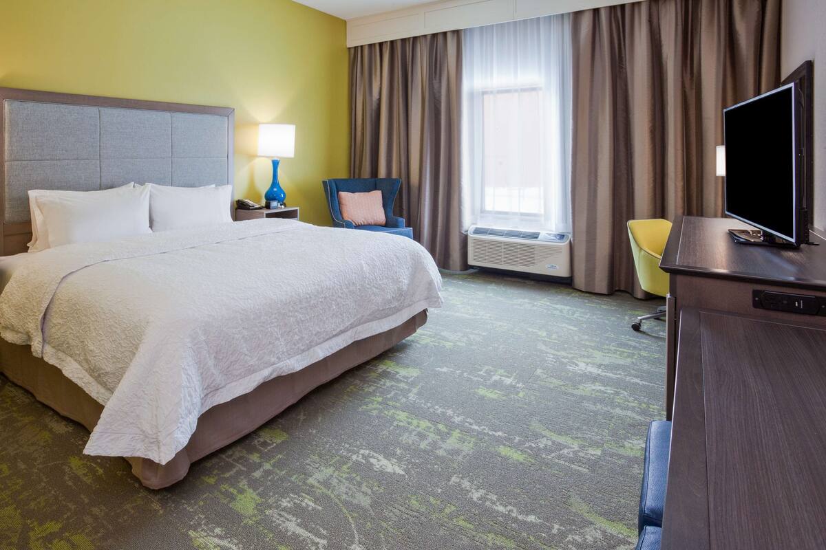 A hotel room with a bed, a desk with a TV, a chair, a lamp, and a window with curtains.