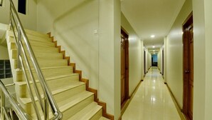 Un pasillo largo y estrecho con puertas de madera a un lado y una escalera con barandillas de metal al otro.