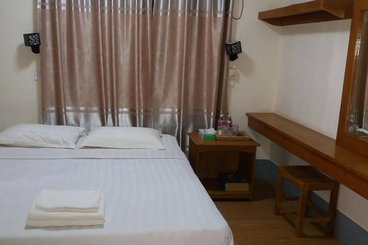A neatly made bed with white linens, a wooden nightstand with a tray holding a bottle and glasses, and a mirror on the wall.