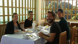 Un grupo de personas cenando en una mesa con mantel blanco, sillas de madera y vistas a la vegetación a través de amplios ventanales.