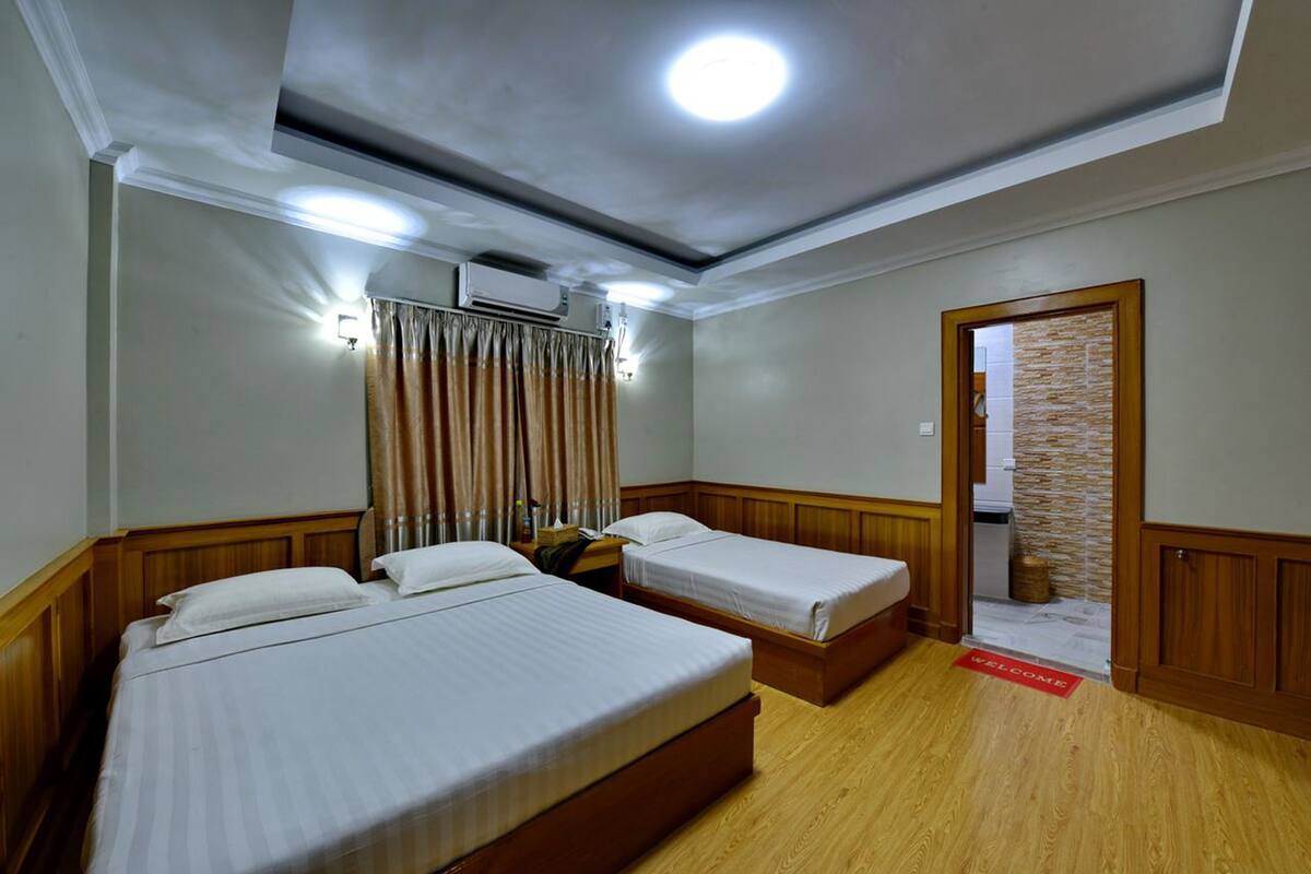 A hotel room with two single beds, a ceiling fan, an air conditioning unit, a wooden floor, and a welcome mat.