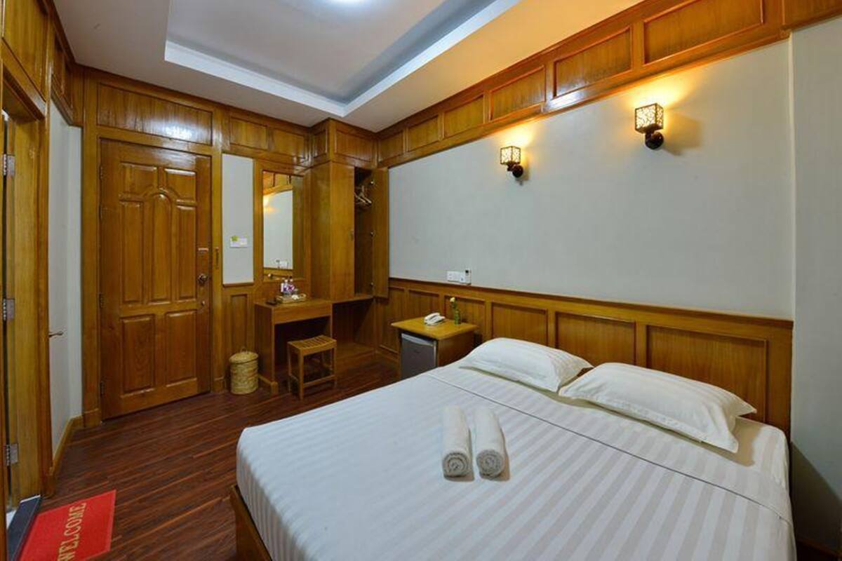 A hotel room with a wooden floor, a bed with white linens, a desk with a chair, a small table with a lamp, and a wall-mounted light fixture.