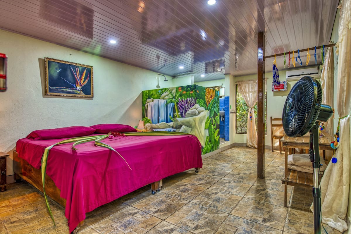 A room with a bed, a shower, a fan, and a mural on the wall.