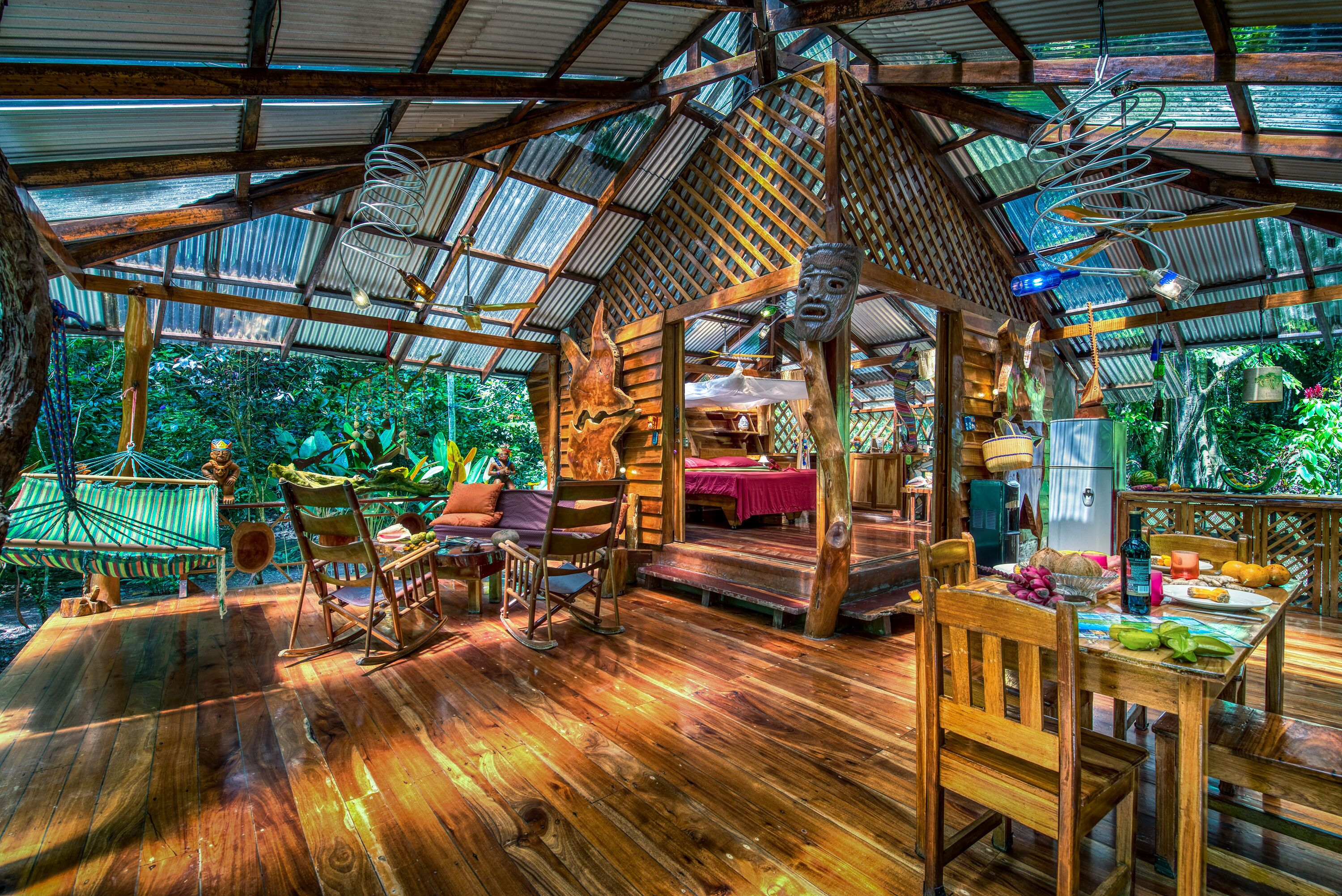 A wooden structure with a thatched roof, a wooden table with fruits, and a hammock.
