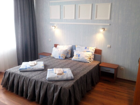 Una cama bien hecha con una colcha gris y dos almohadas, flanqueada por mesitas de noche con lámparas, en una habitación con paredes azul claro y suelo de madera.
