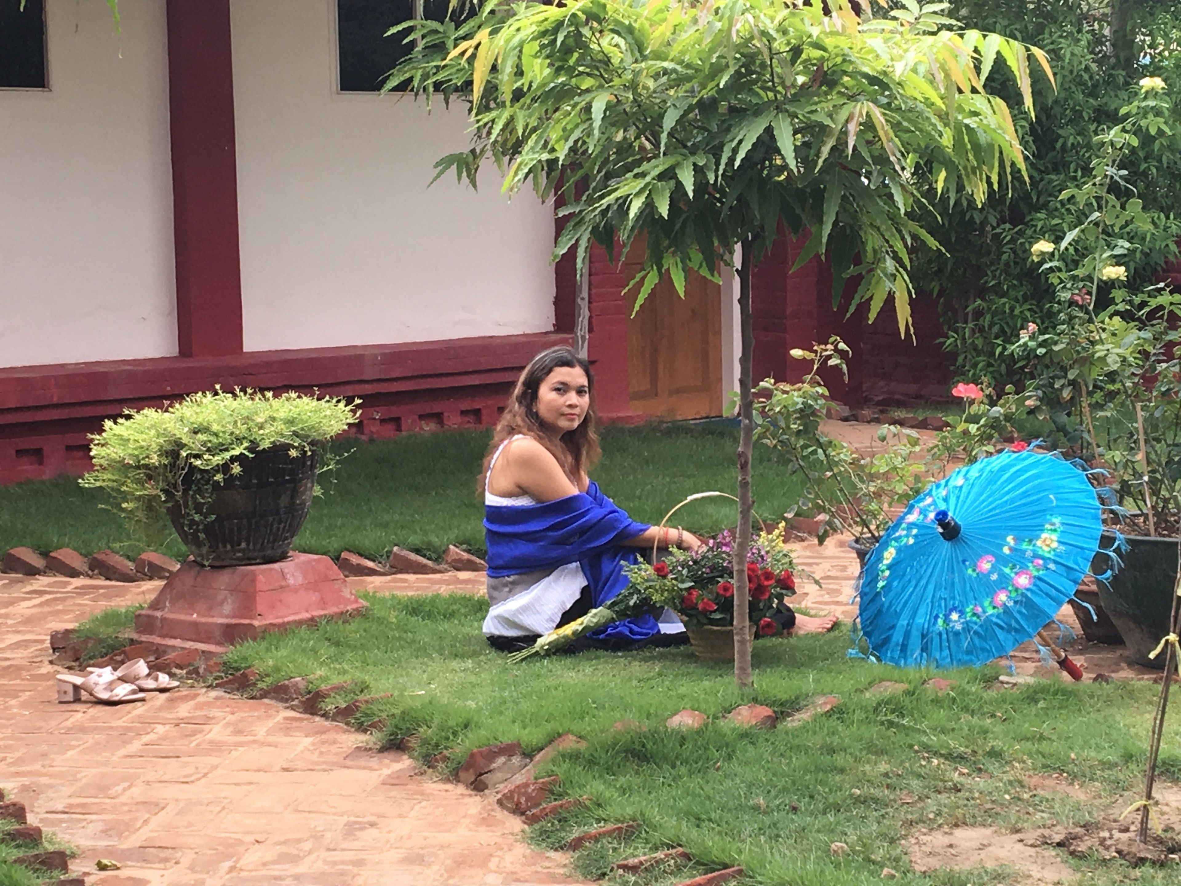 Une personne est assise sur un chemin de briques dans un jardin, entourée de plantes en pot et d’un parasol bleu.