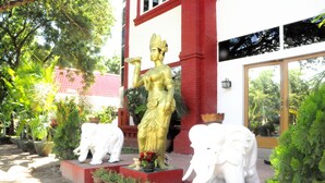A statue of a deity on a pedestal, flanked by two white elephant statues, in front of a building with red pillars and large windows. - La Casa Di Bagan Nan Eain Thu (Nyaung-U)
