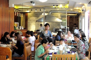 A restaurant with wooden furniture, a kitchen area, and a group of people dining. - La Casa Di Bagan Nan Eain Thu (Nyaung-U)