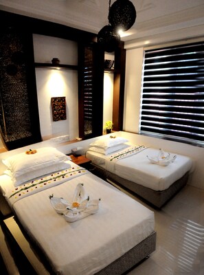 A hotel room with two beds, each with white linens and floral decorations. There is a large window with blinds, a wall-mounted light fixture, and a decorative wall piece. - La Casa Di Bagan Nan Eain Thu (Nyaung-U)