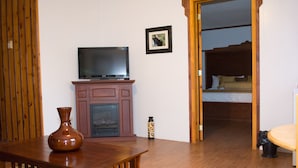 Una habitación con una mesa de madera, un televisor sobre un mueble, una chimenea, una cama con cabecero de madera y una puerta que da a otra habitación.