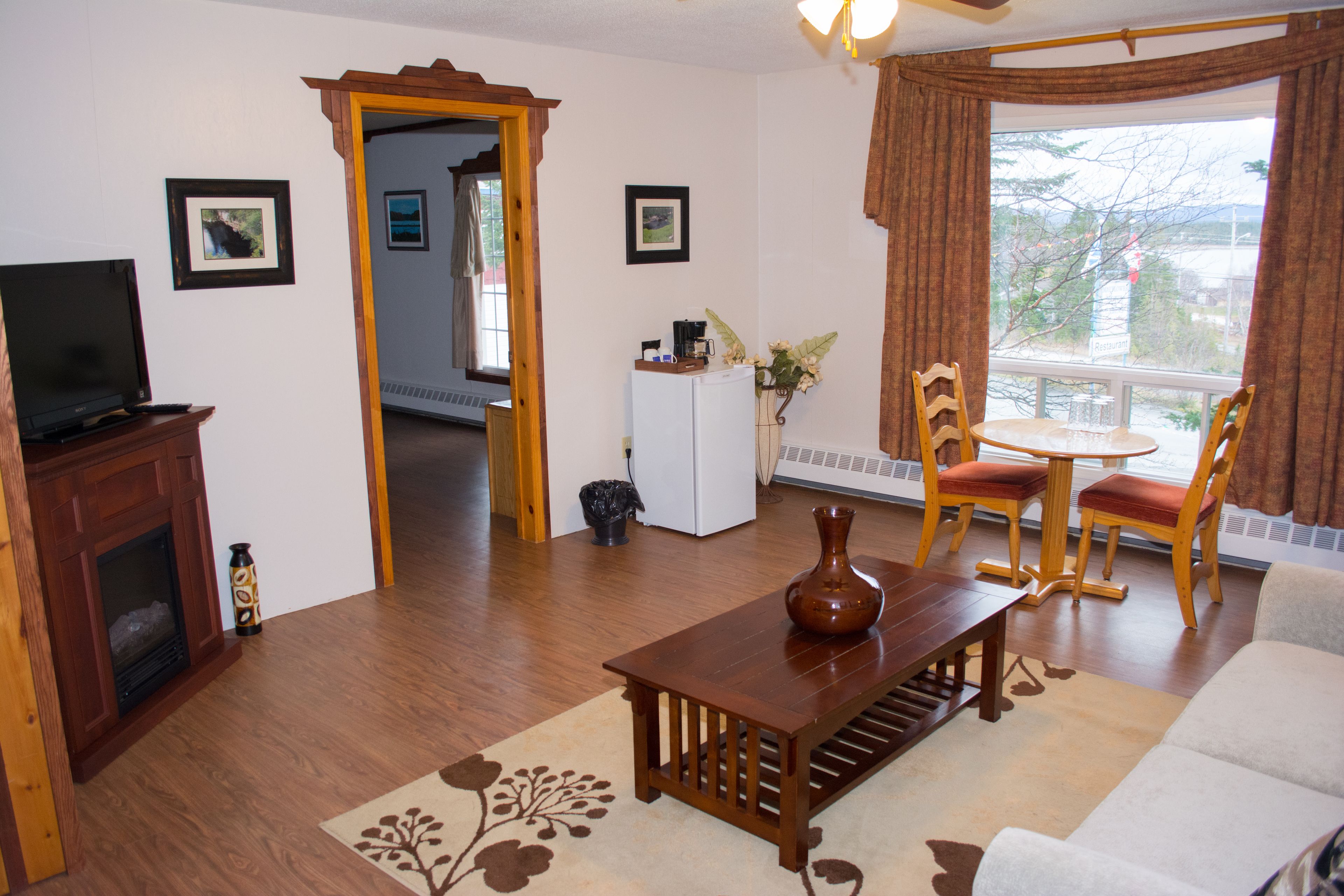 A living room with a fireplace, a television, a coffee table, and a sofa. There is a window with curtains, a door, and a small table with a vase.