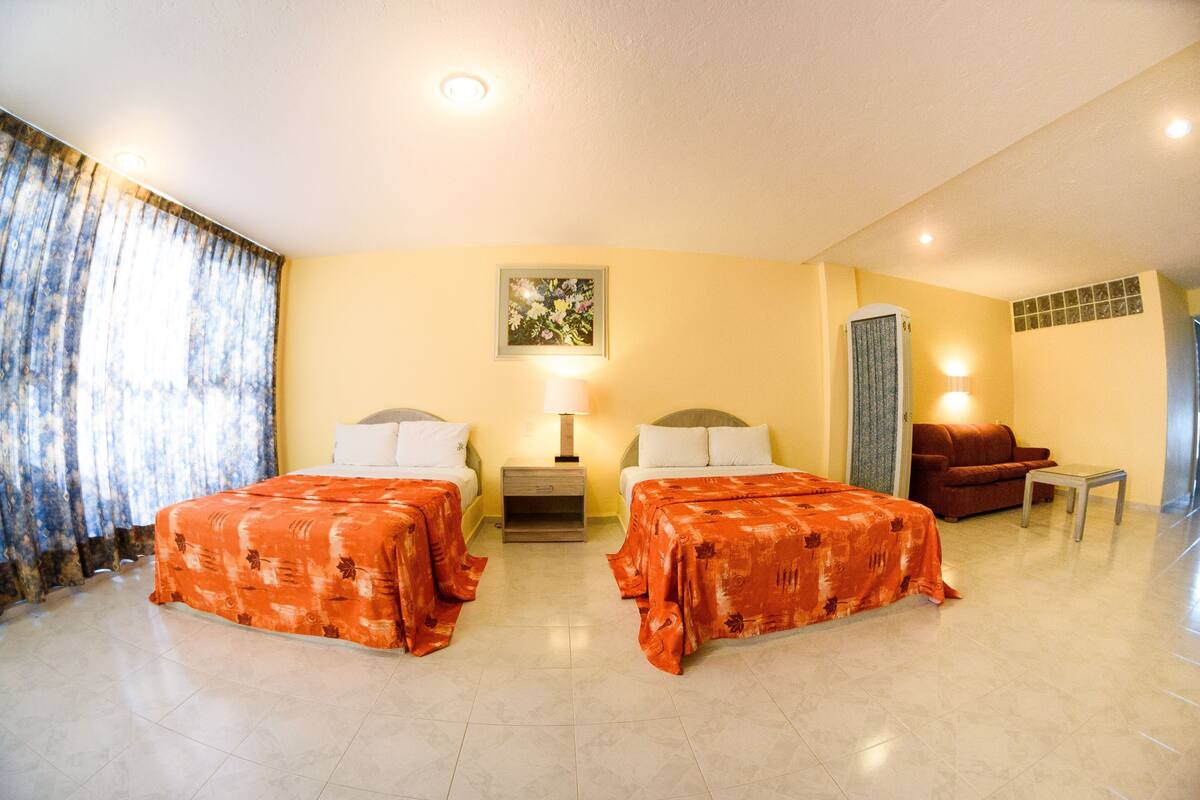 Ein Hotelzimmer mit zwei Betten, einem Schlafsofa, einem kleinen Tisch und einem Nachttisch mit Lampe.