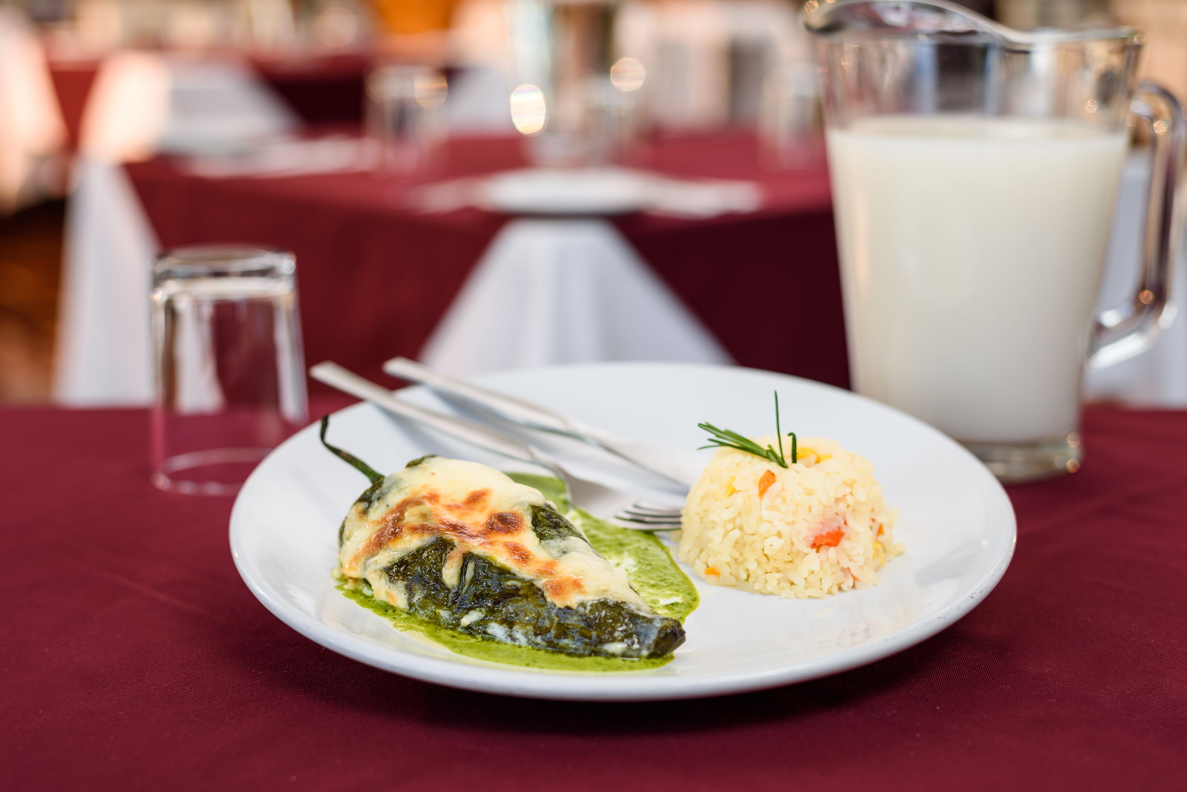 A plate with a stuffed vegetable and rice, a glass of milk, and a pitcher on a table with a red tablecloth.