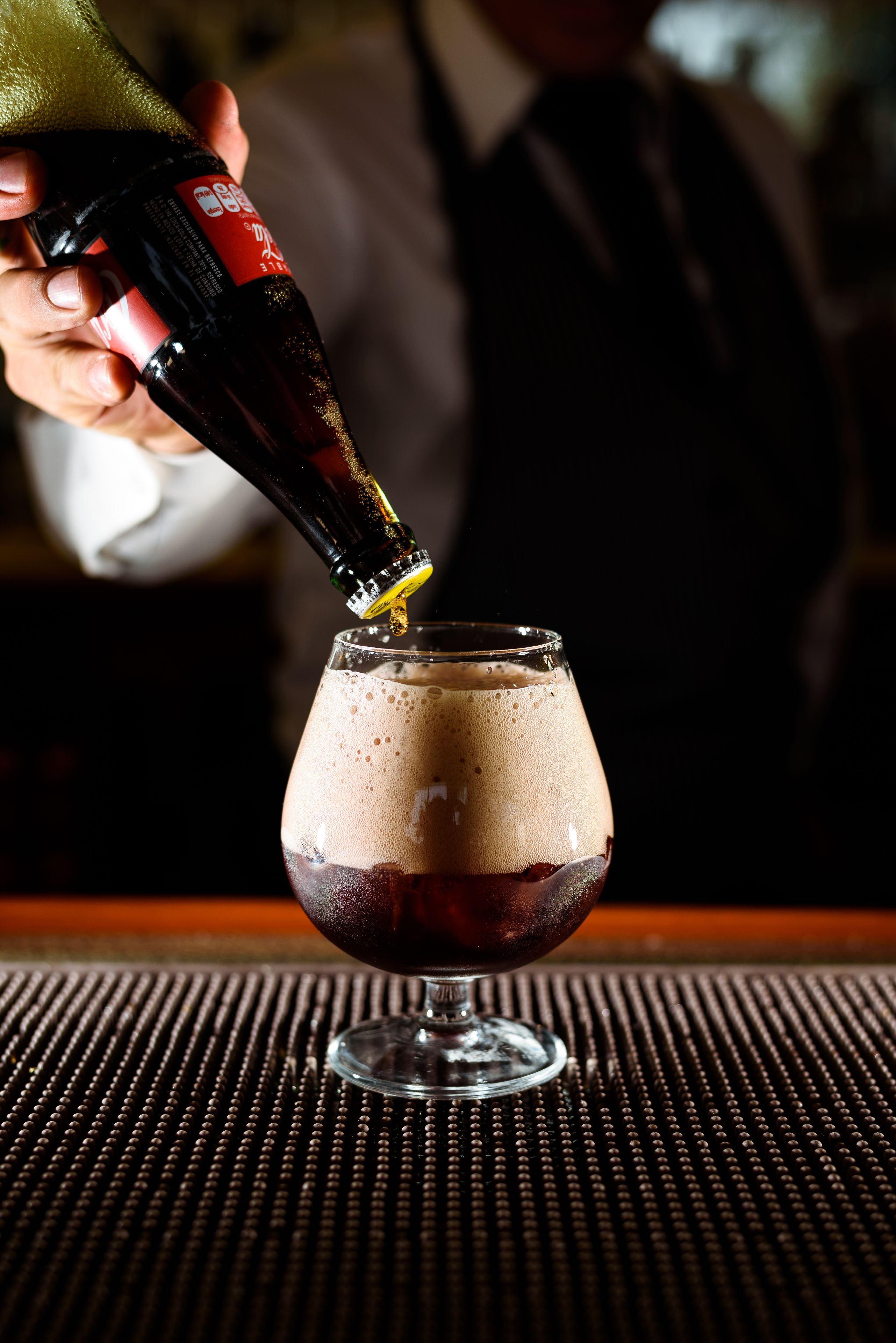 A hand pouring a dark liquid into a glass with a foamy head, placed on a textured surface.