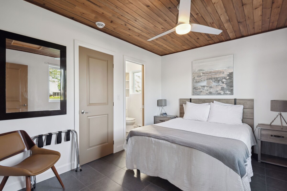 Une chambre à coucher comprenant un lit, une chaise, une table de chevet, une lampe, un ventilateur de plafond et un tableau accroché au mur.