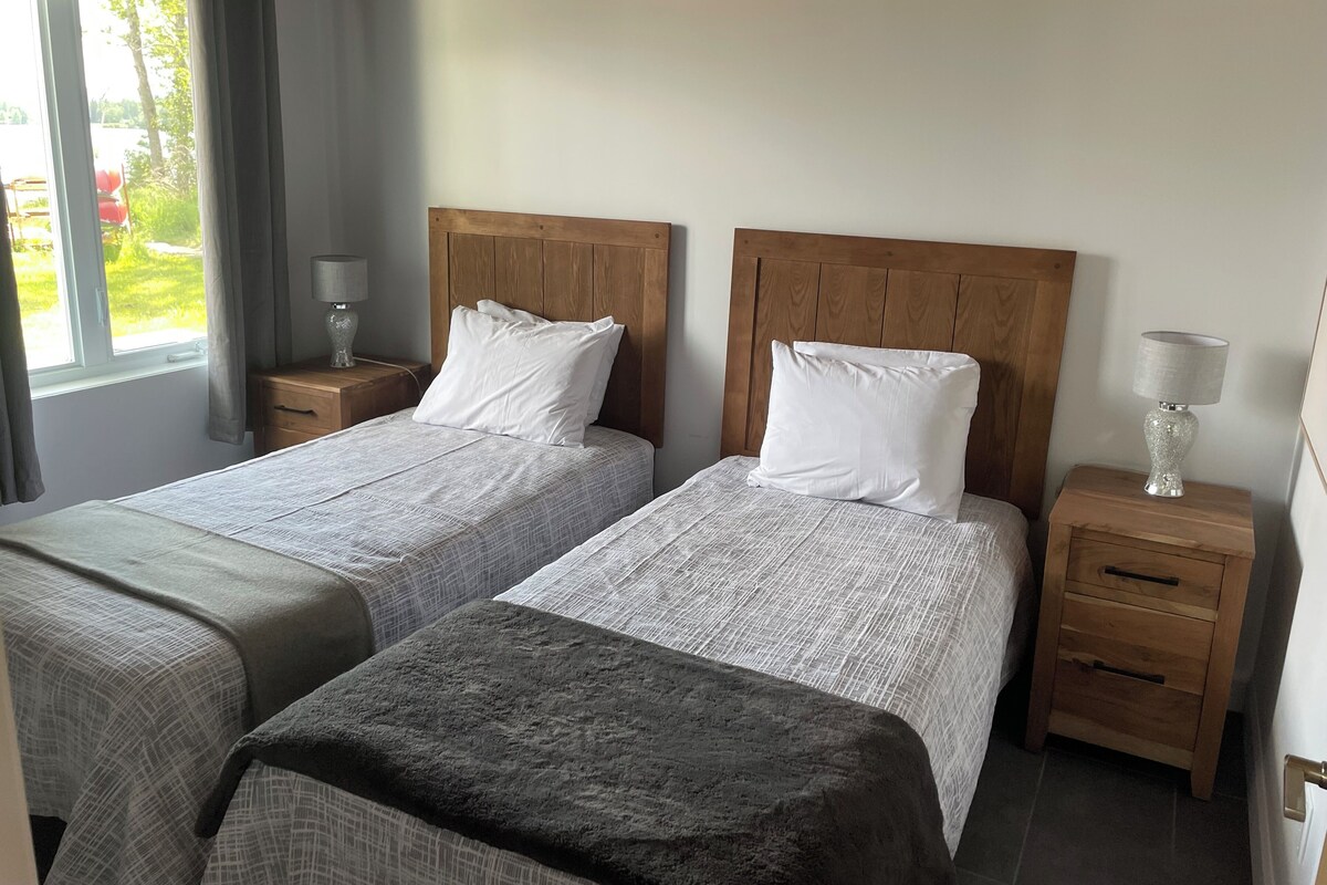 Une chambre double avec deux lits, des têtes de lit en bois, des tables de chevet avec des lampes et une vue sur la verdure environnante.