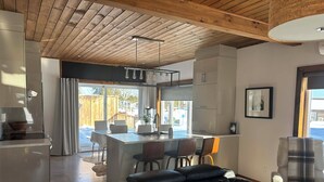 A modern kitchen with a central island, dining area with chairs, a black leather sofa, and a large window. - Les Chalets Baie Cascouia (Larouche)
