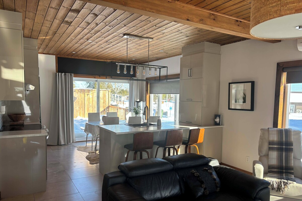 Une cuisine moderne avec un îlot central, un coin repas avec des chaises, un canapé en cuir noir et une grande fenêtre.