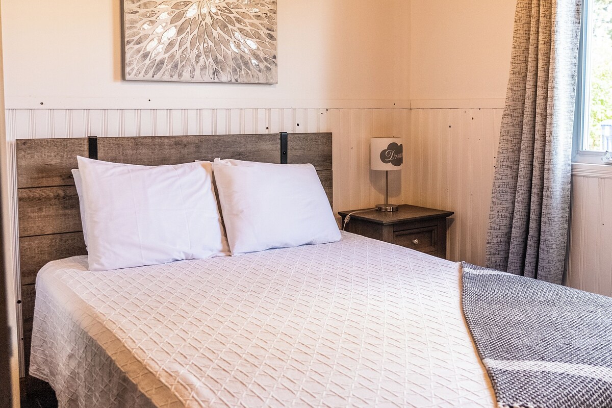 Une chambre avec un plafond en bois, un lit recouvert d’une courtepointe matelassée, une table de chevet avec une lampe et une fenêtre avec des rideaux.