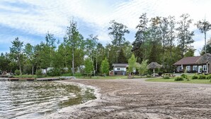 A lakeside property with a sandy beach, a wooden dock, and a house with a thatched roof. - Les Chalets Baie Cascouia (Larouche)