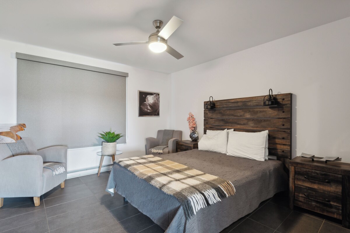 Une chambre à coucher comprenant un lit, une chaise, une table de chevet, un ventilateur de plafond et une fenêtre avec des stores.