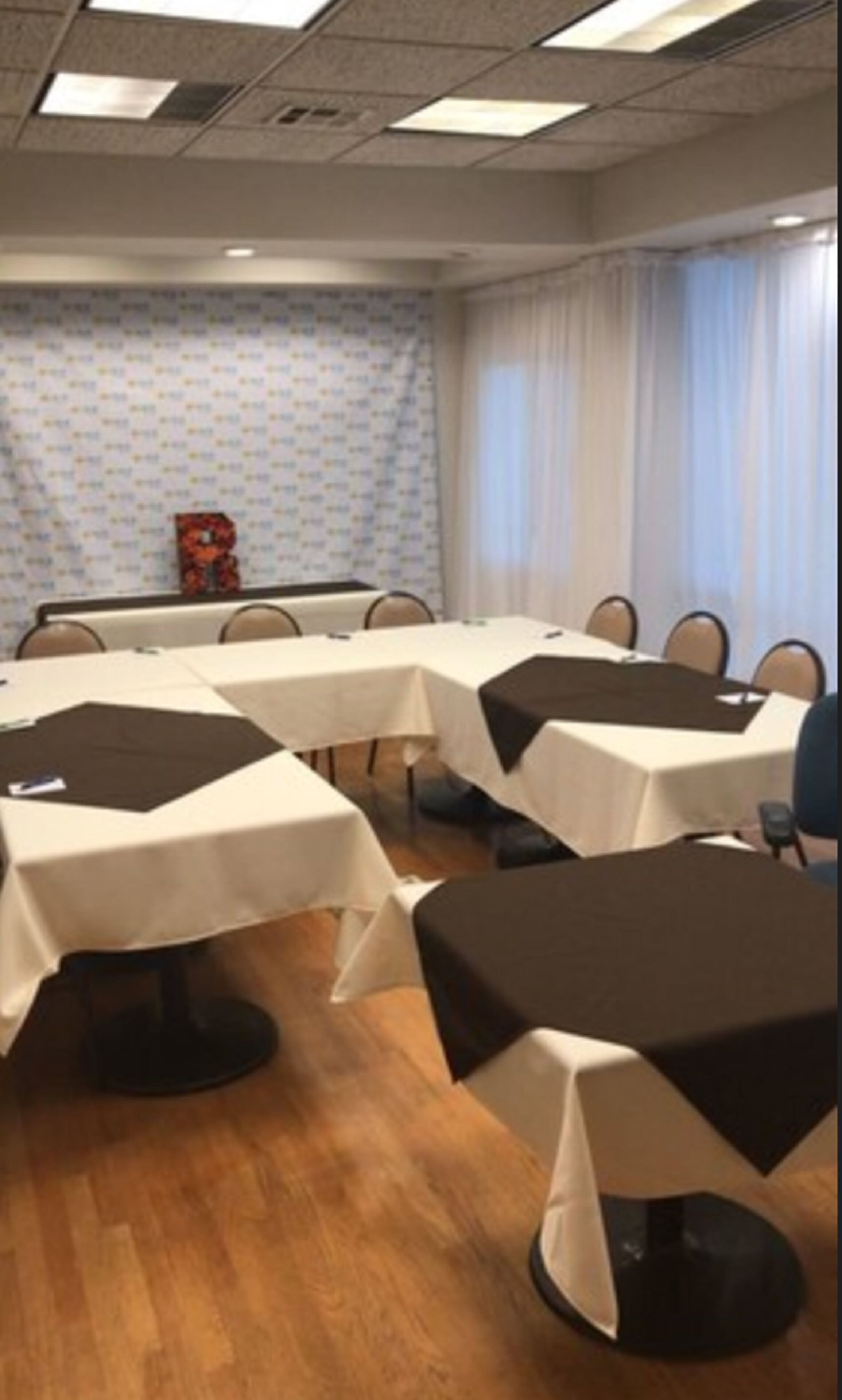 Ein Konferenzraum mit mehreren Tischen, die jeweils mit einer weißen Tischdecke und einer schwarzen Tischläufer bedeckt sind. Der Raum hat einen Holzboden und eine Wand mit Muster.