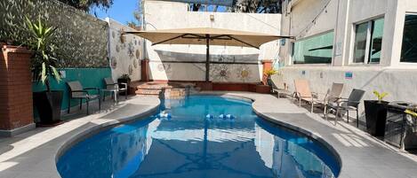 A swimming pool with a sunshade, surrounded by lounge chairs and a building with large windows.