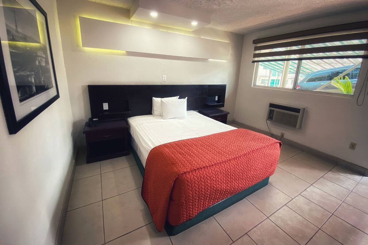 Una habitación de hotel moderna con una cama grande, una mesita de noche, un televisor de pared y una ventana con persianas.