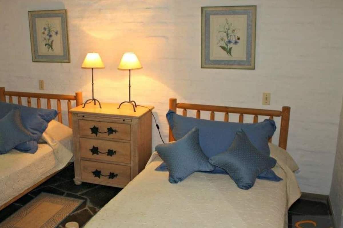 Una habitación con dos camas individuales, una mesita de noche de madera con dos lámparas, y dos cuadros con motivos florales en la pared.