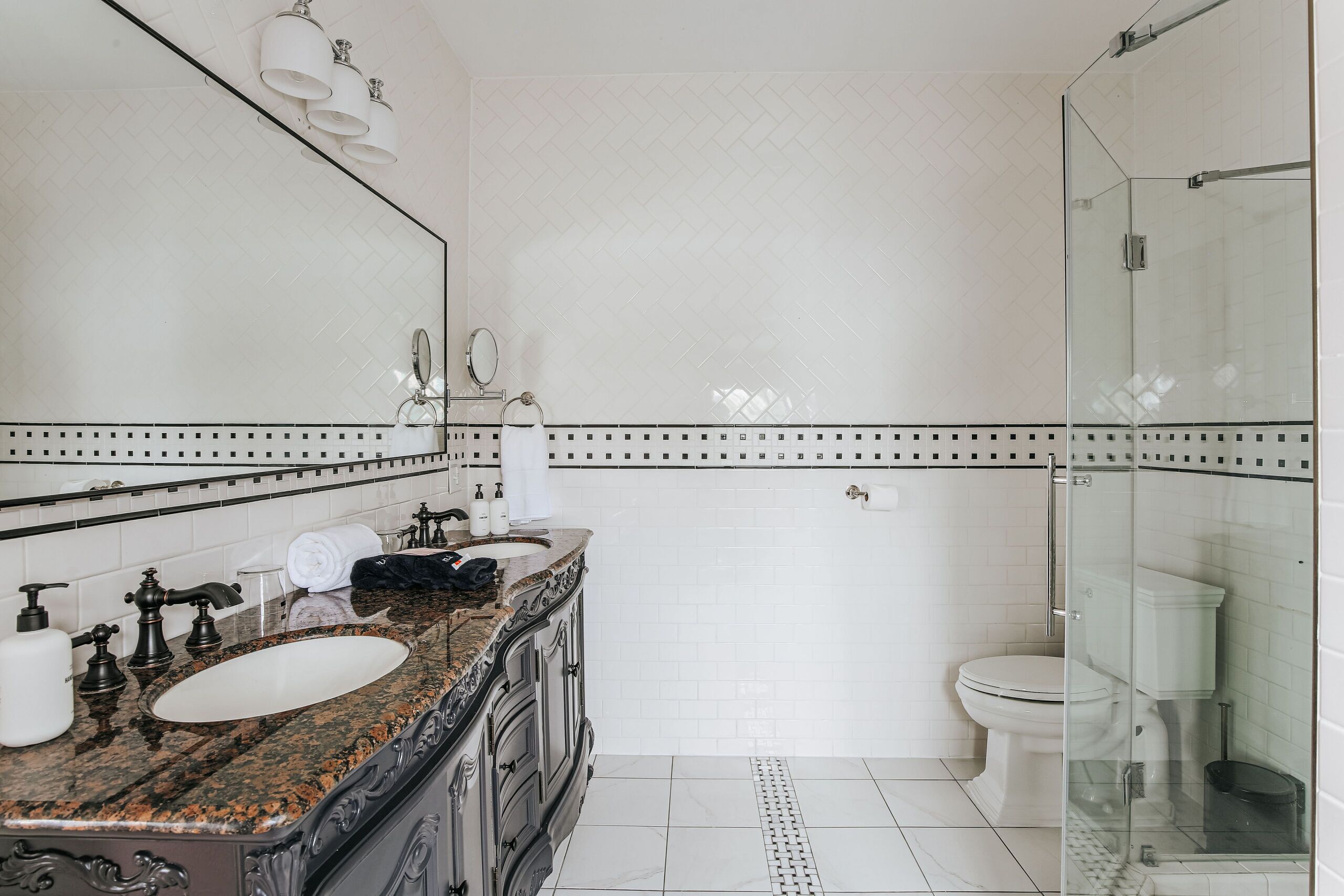 A bathroom with a double sink, a mirror, a toilet, and a glass shower enclosure.