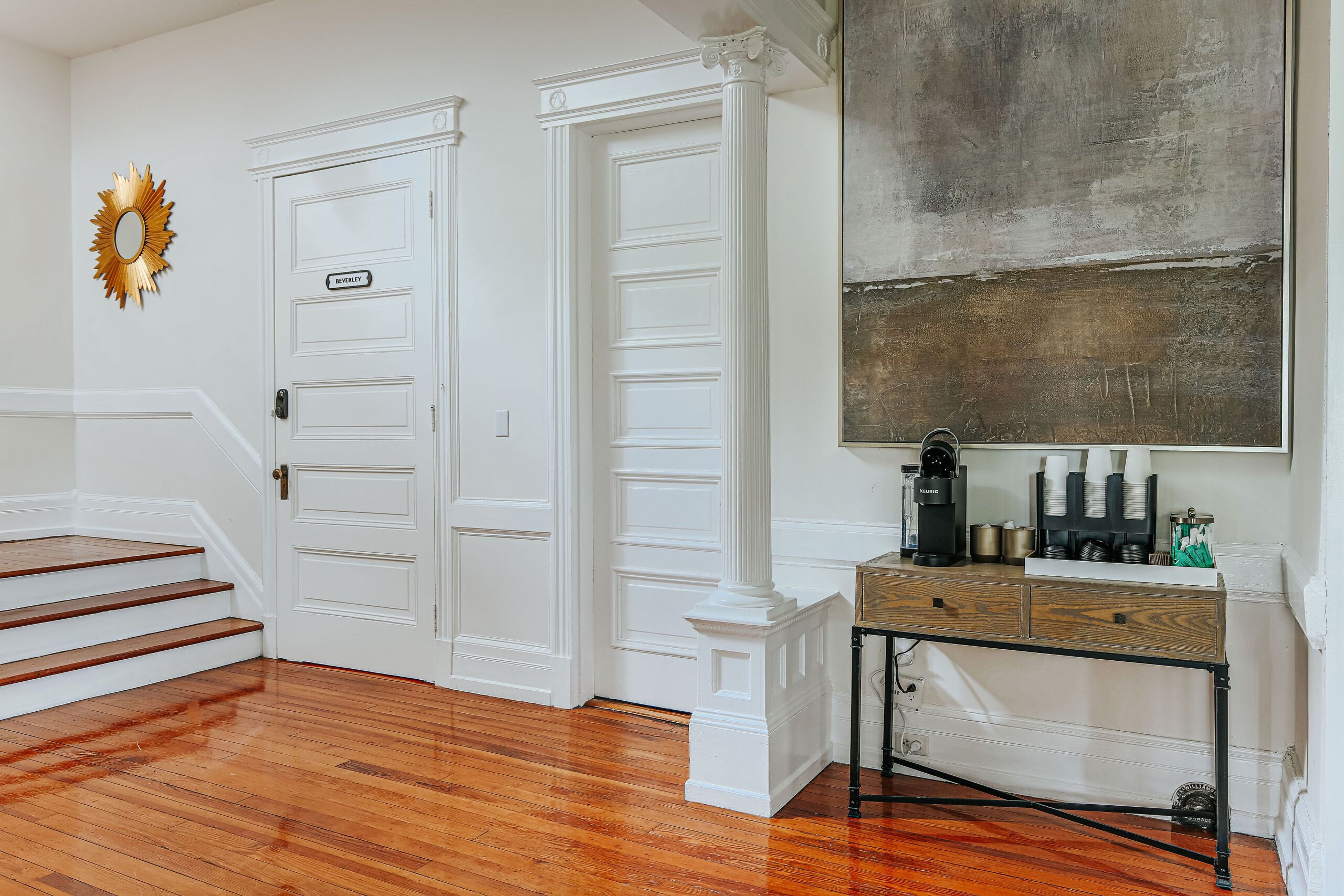 A hallway with white doors, a wooden staircase, and a coffee station with a large painting above.