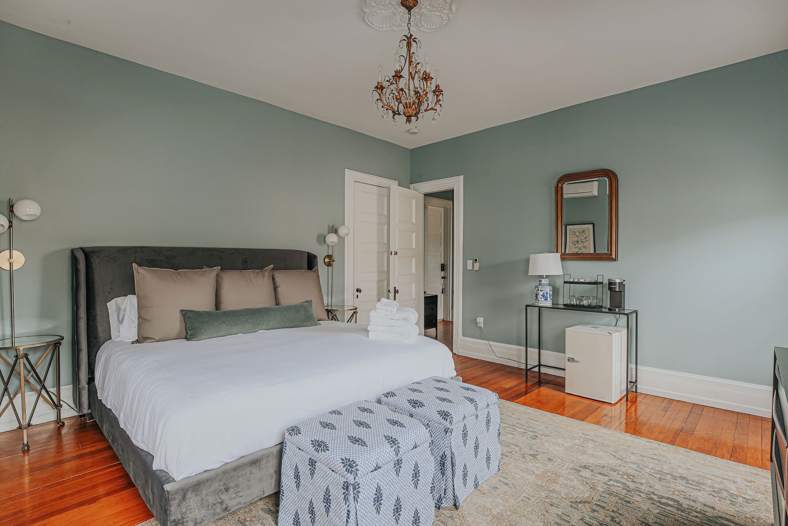 A bedroom with a large bed, bedside tables, a dresser, a mirror, and a door leading to another room.