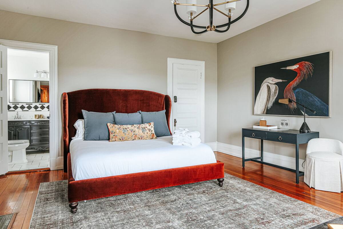 A bedroom with a red velvet bed, grey and blue pillows, a grey rug, a black desk, and a white chair.