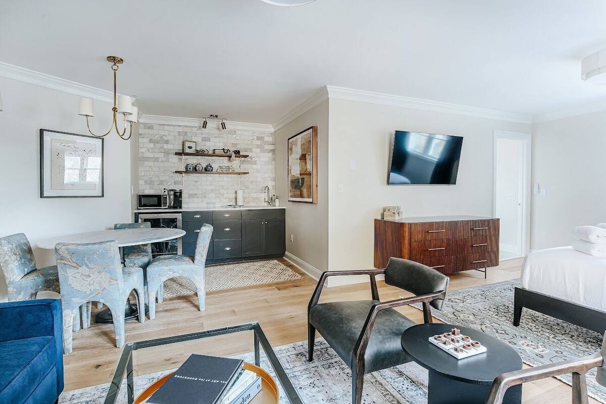 A modern hotel room with a dining area, kitchenette, and a TV mounted on the wall.